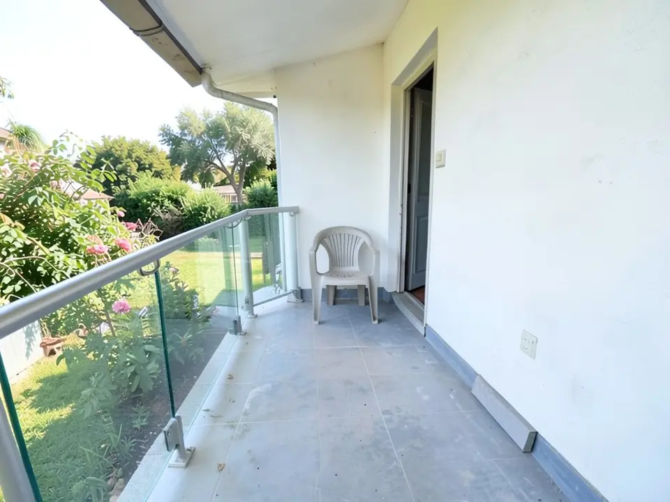 The same balcony with a clear glass-panel railing and a thin steel handrail along the left side, a lighter grey floor with faint tile joints, the same white wall, door, plastic chair and garden view