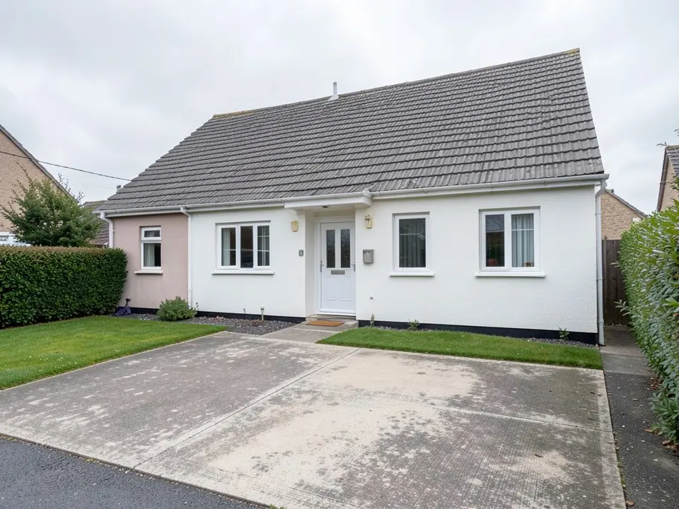 Front of a detached single-storey white rendered house with a grey tiled roof, a white front door with two windows on its right and one on its left, a pink-painted side wing on the left, a small lawn, a hedge and a wide concrete driveway in front