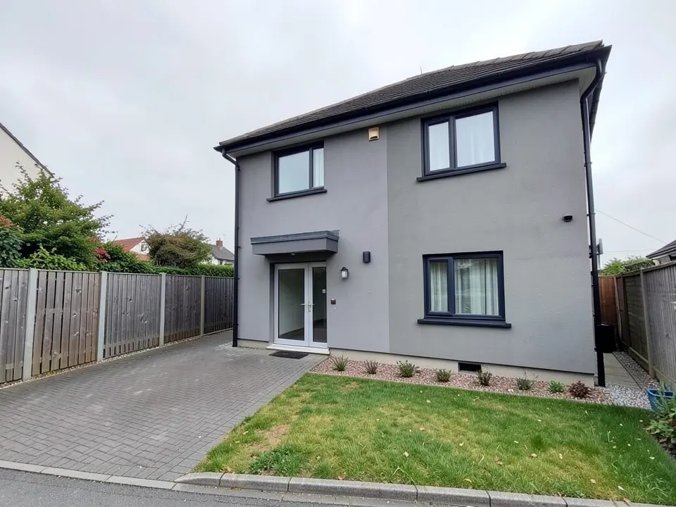 Side-on view of a two-storey grey rendered house with dark window frames and a small flat canopy over a glazed door, a block-paved driveway running along the left side beside a wooden fence, a small lawn and the pavement edge