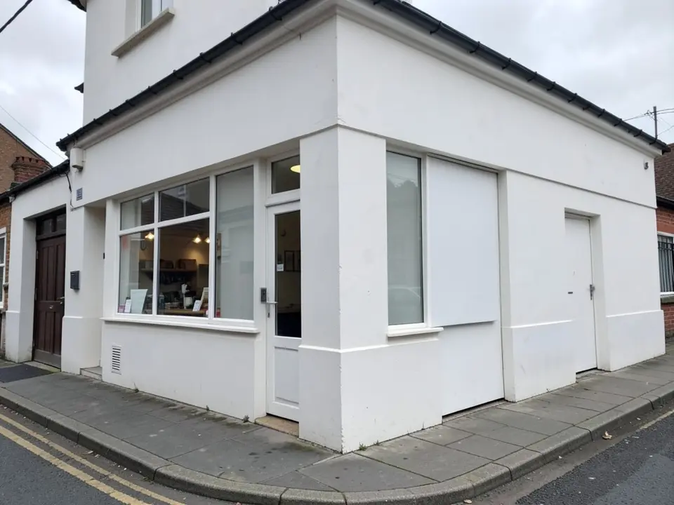 A white rendered corner building with a ground-floor window and a white door, a blank window panel to the right, a dark maroon door at the far left, a black gutter, a paved pavement and a street with double yellow lines