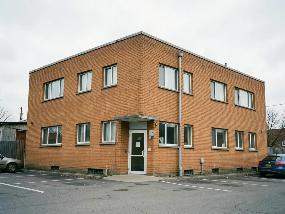 A two-storey office building with orange brick walls, small white-framed windows, a small canopy over a white entrance door, a drainpipe, and a car park with white bay lines and two parked cars