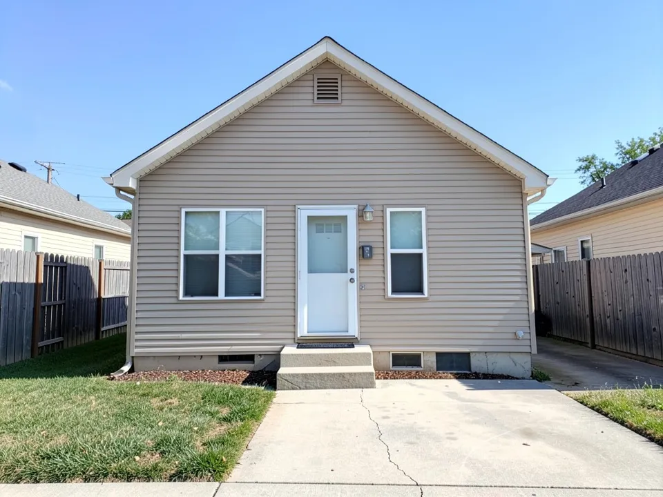 A plain one-storey house with beige vinyl siding and a front gable, a white door, two sliding windows, a concrete step, a lawn, a driveway and fences