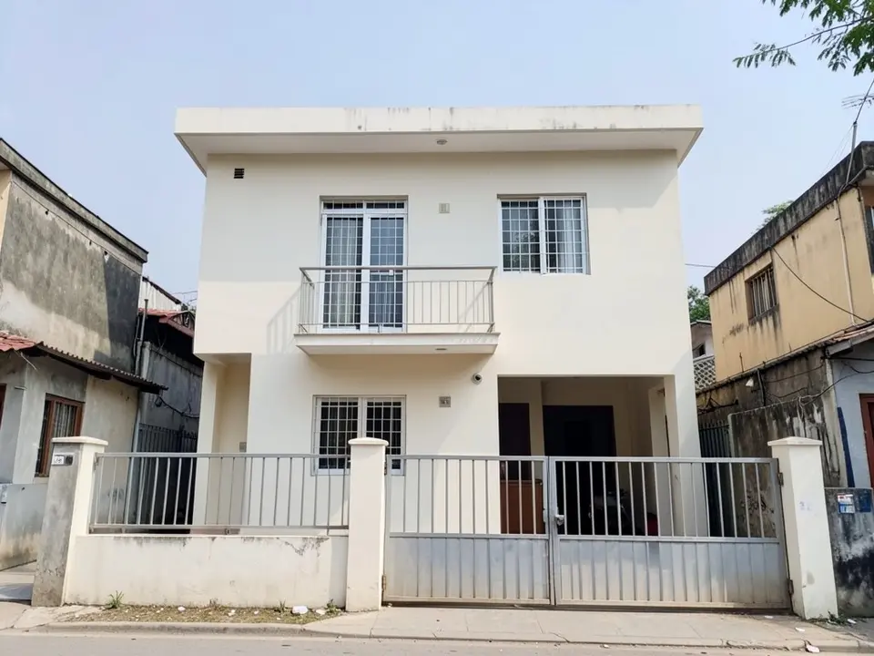 Front of a plain two-storey off-white house on a narrow city plot, with a flat roof, a small first-floor balcony with a metal railing, windows with metal grills, a car porch and a metal gate in a low boundary wall
