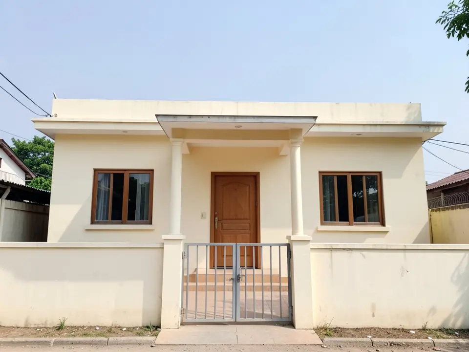 Front of a single-storey cream house with a flat roof, two wood-framed windows, a wooden front door and a small verandah with a thin slab roof on two slim square pillars, behind a low boundary wall with a metal gate