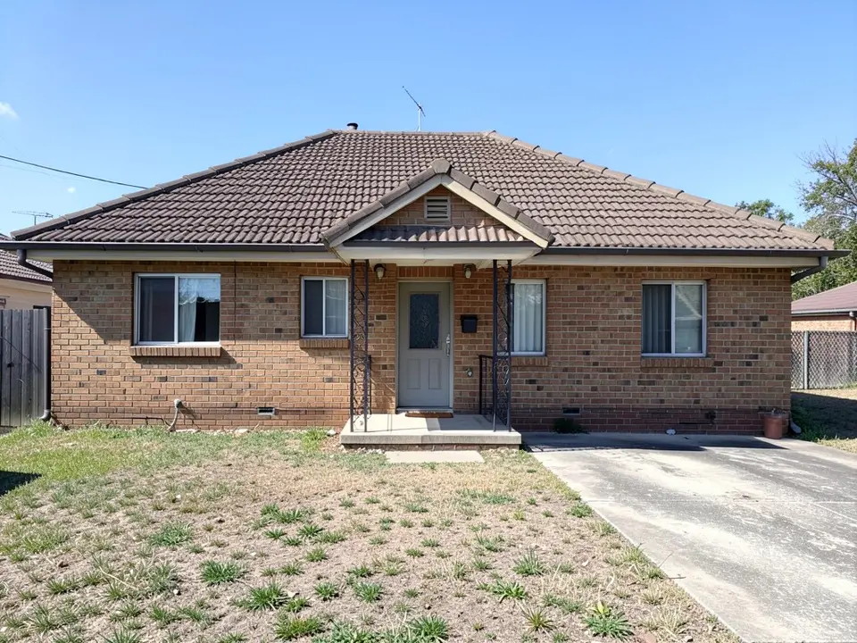 Front of a dated single-storey brown brick bungalow with a hipped brown tile roof, small windows, a narrow porch with wrought-iron posts under a small gable, a concrete driveway on the right and a patchy lawn