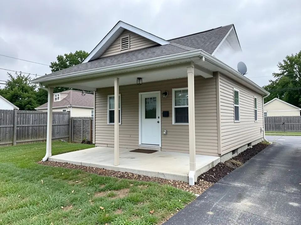 Front of a small beige-sided house with a covered porch held up by three thin cream posts, a white ceiling, a bare concrete floor, no railing, a white front door, a lawn and a dark driveway