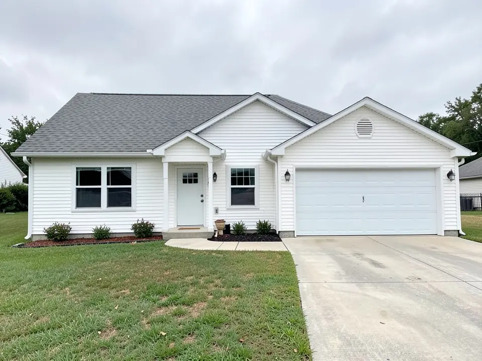 Front of a single-storey white-siding ranch house with a grey shingle roof, a pale green front door under a small gabled porch, two windows, an attached double garage on the right with a white garage door and a vent in its gable, a concrete driveway and a lawn