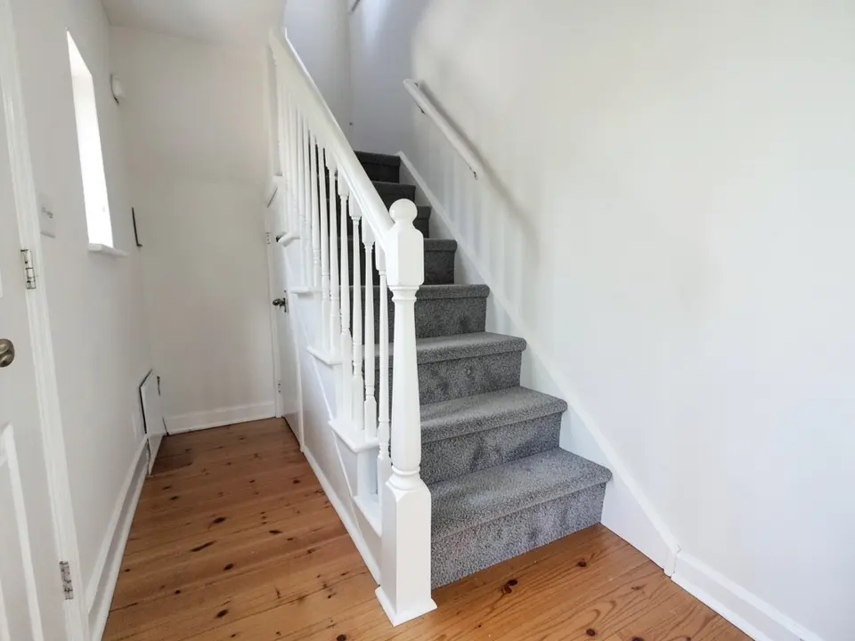 A narrow staircase with a grey carpet on each step, white risers, white spindles, a white newel post with a ball top and a white wall handrail, white walls and a pine plank floor