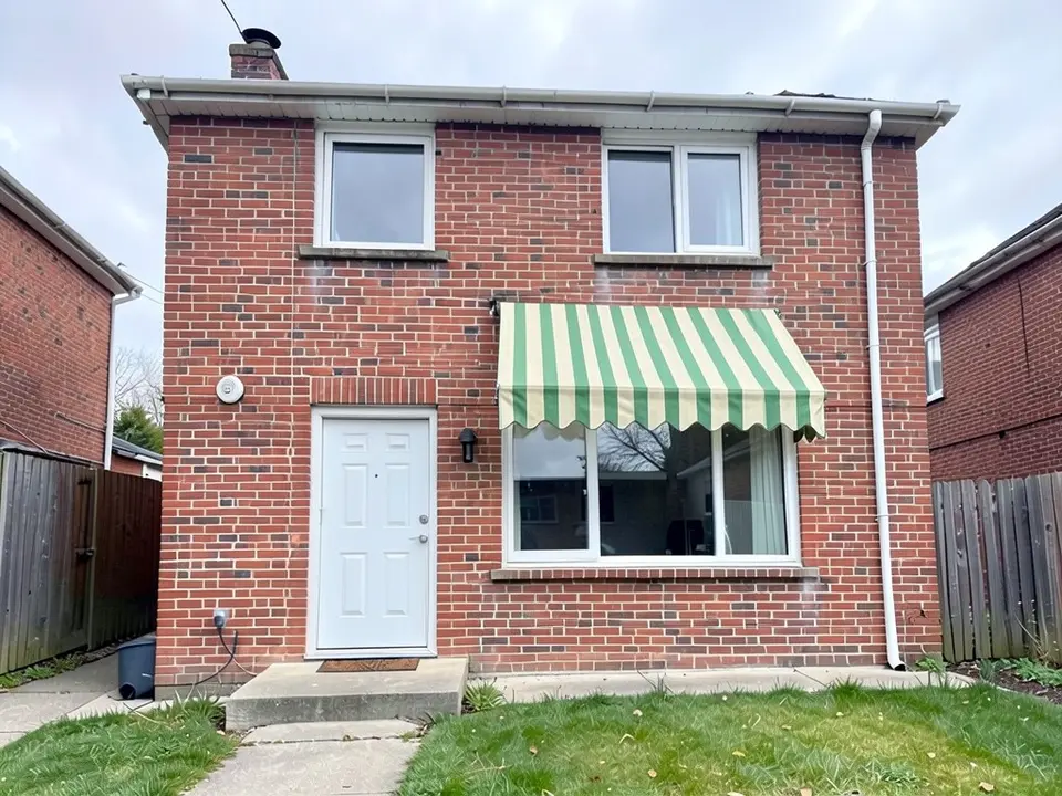 The same house with a green and cream striped fabric awning with a scalloped edge hanging over the top of the large ground-floor window, with the same door, step, upstairs windows, downpipe, fences and lawn