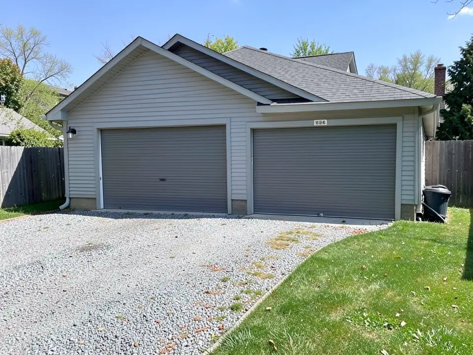 Side of a detached two-car garage with grey siding and two grey roll-up doors, a pitched roof, a gravel drive, a wooden fence and a small lawn