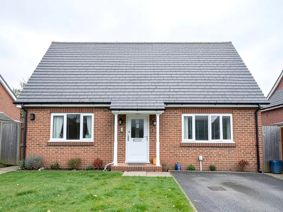 Front of a detached brick bungalow with a plain grey pitched roof without roof windows, a white front door with a small canopy, a window on each side, a lawn and a driveway