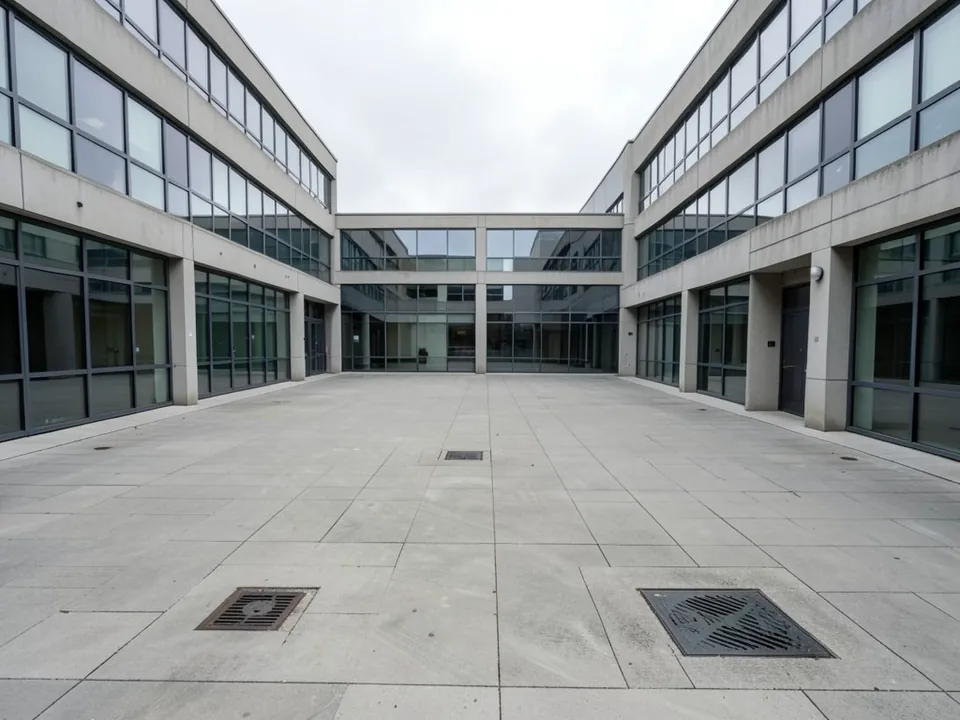 Photo of an empty courtyard between low concrete and glass office buildings: flat grey paving slabs, two large drain covers in the foreground and smaller ones further back, no plants or furniture, overcast sky