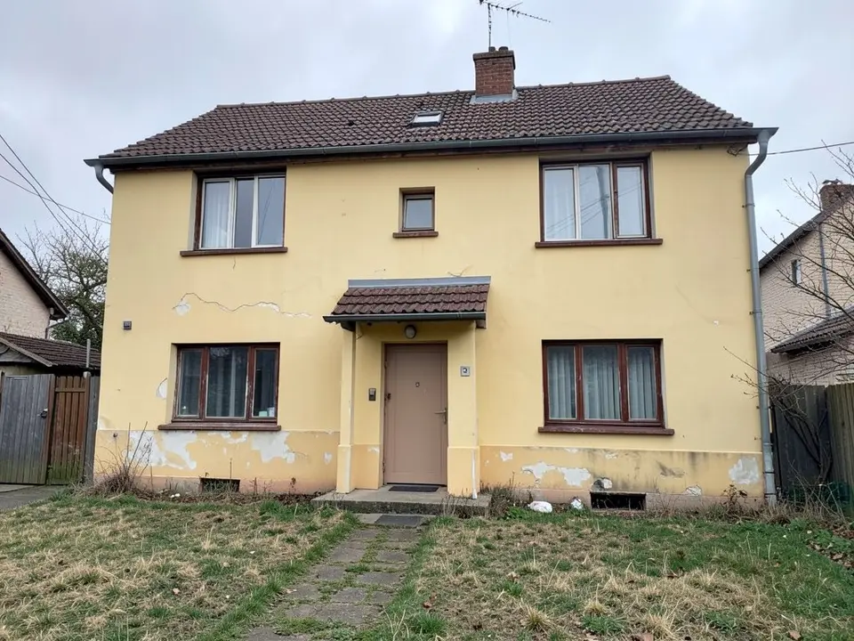 Photo-style front view of a two-storey house with faded yellow walls with cracked and flaking patches, a brown tiled roof with a chimney, brown window frames, a small porch over a pale brown door and a patchy lawn with a stone path